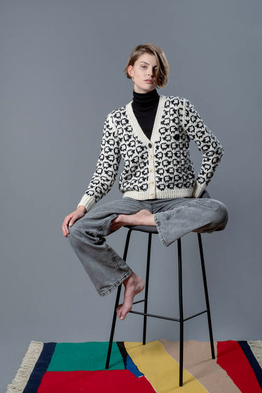 Woman sitting on a chair wearing a wool cardigan with a turtleneck underneath. The cardigan has a herd of white sheep all over with a single black sheep on the left side.